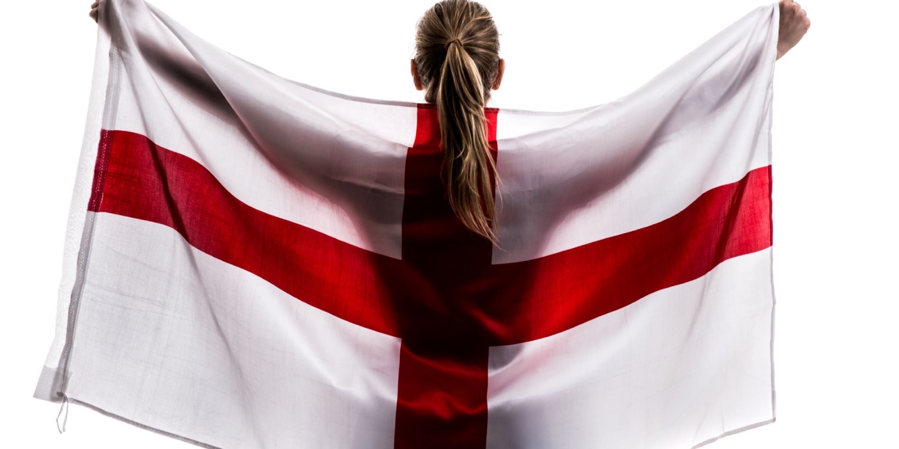 A woman with long blond hair with her back to the camera and arms raised is holding an England flag behind her